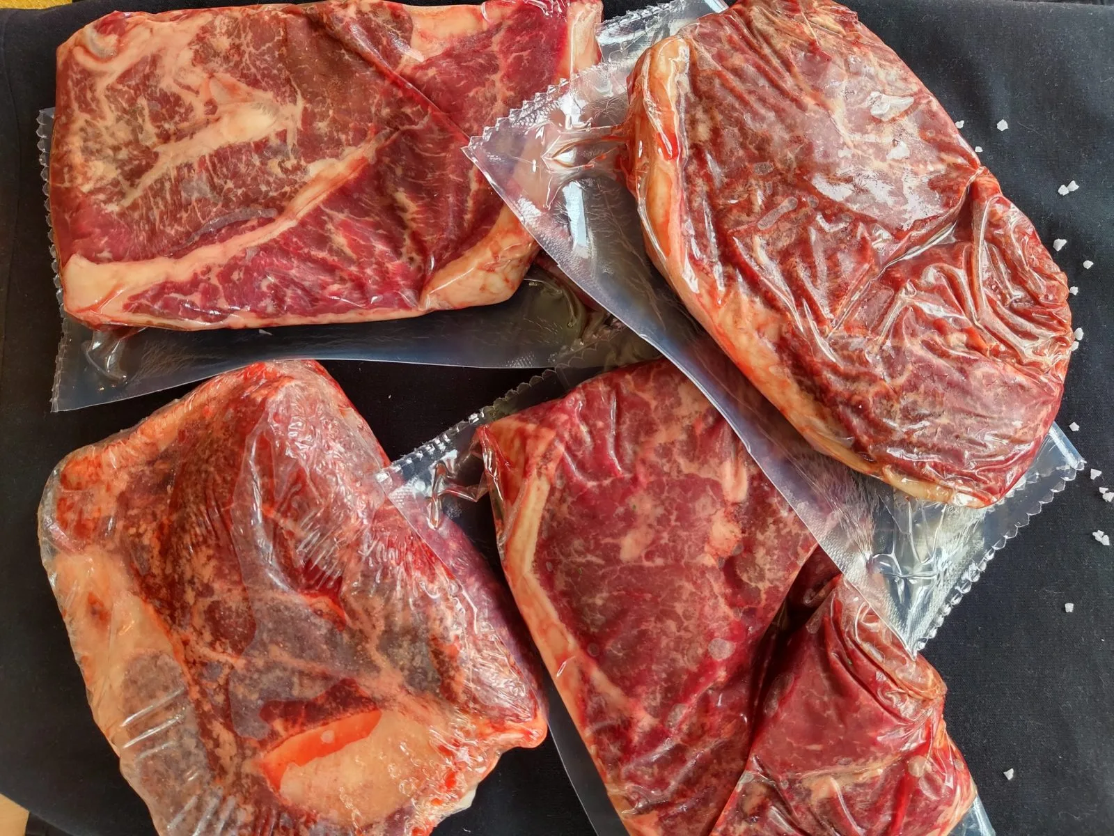 Five vacuum-sealed, well-marbled Nadig Farms beef steaks laid out on a black surface with coarse salt flakes scattered around them
