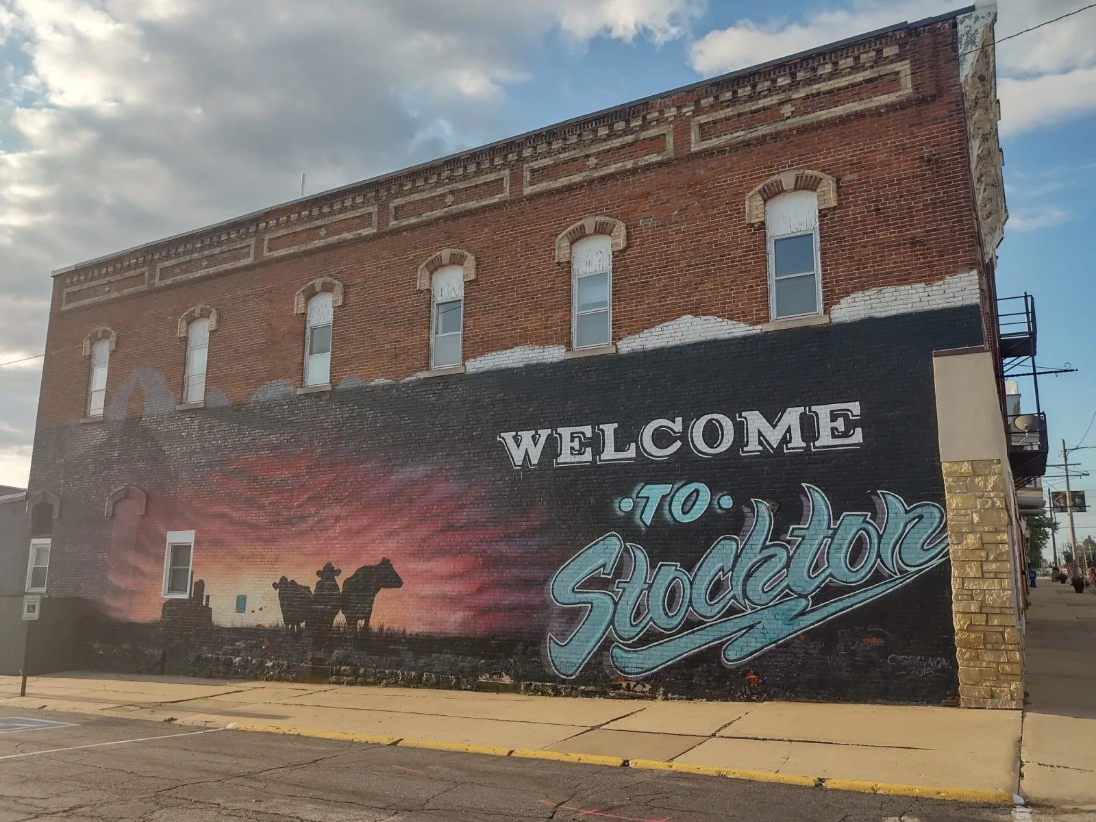 "Welcome to Stockton" mural on a brick building in Stockton, Illinois, with cattle silhouetted against a sunset