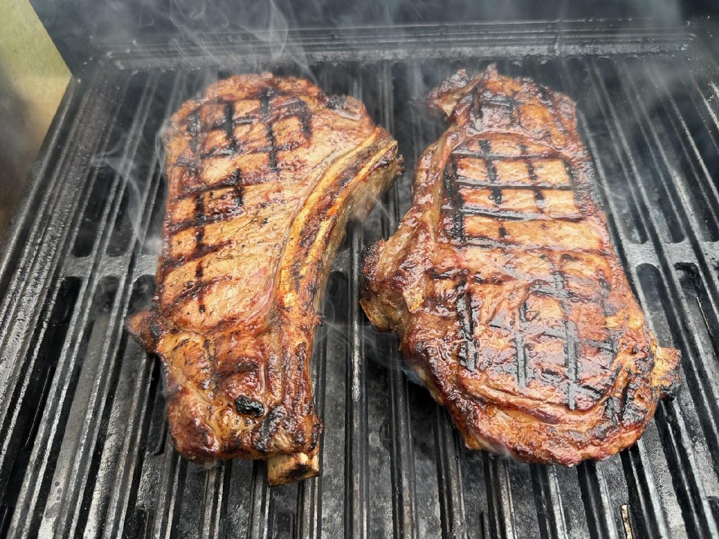 Ribeye steaks searing over open flame with smoke rising off the grill — grain-finished beef from Nadig Farms.