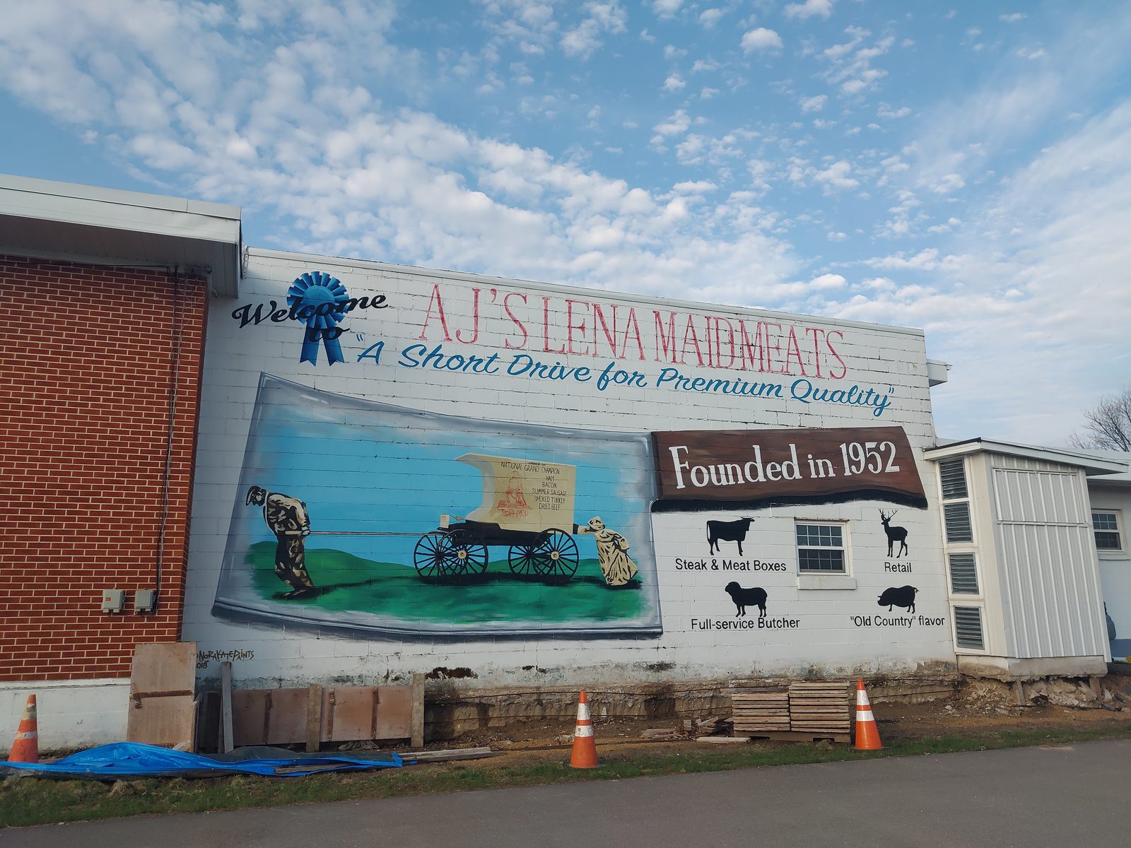 Painted welcome mural on the exterior of AJ's Lena Maid Meats in Lena, Illinois, reading "Founded in 1952," with icons for steak and meat boxes, retail, full-service butchering, and Old Country flavor.